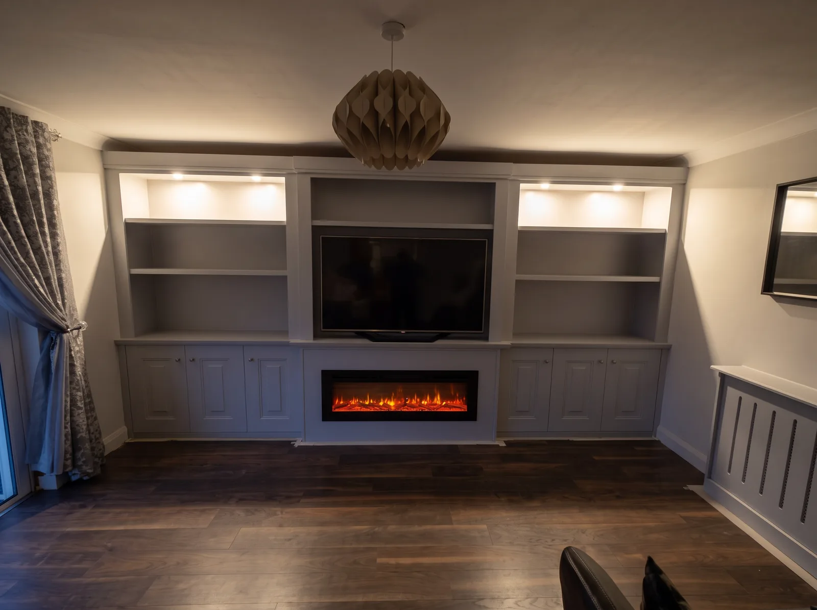 Pale grey media wall at dusk with downlit shelving, inset fire, panelled cupboards and a matching radiator cover